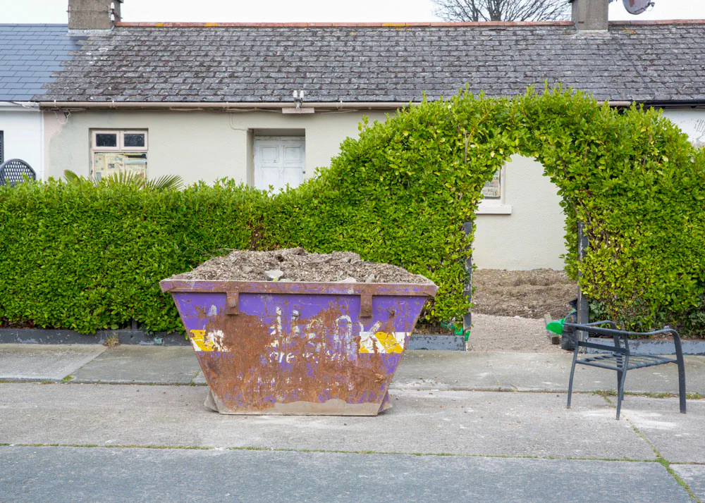 Purple skip on footpath in front of luscious green hedge
